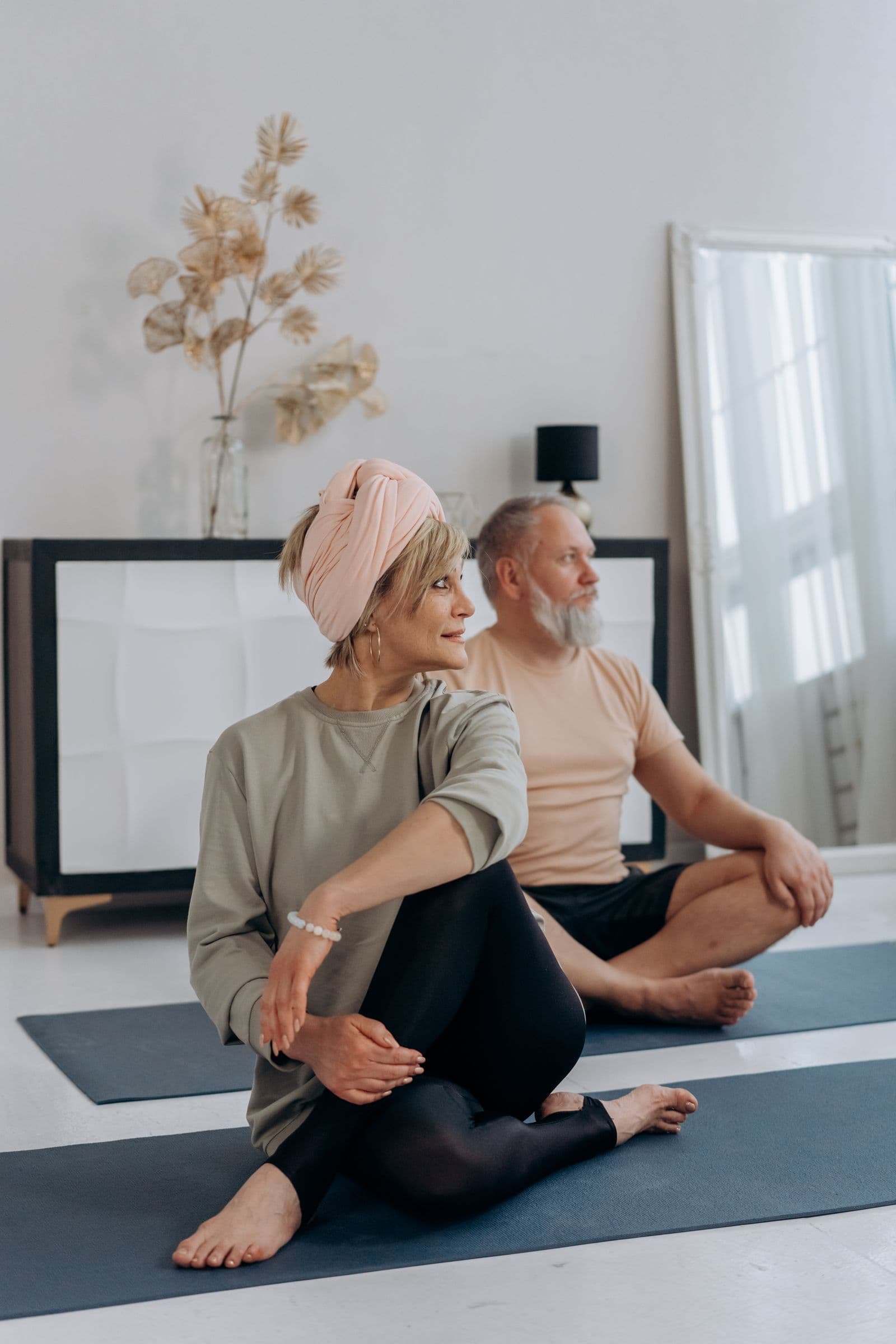 Two middle-aged adults sitting on yoga mats on the floor of a bright living room, one in a seated spinal twist and one cross-legged
