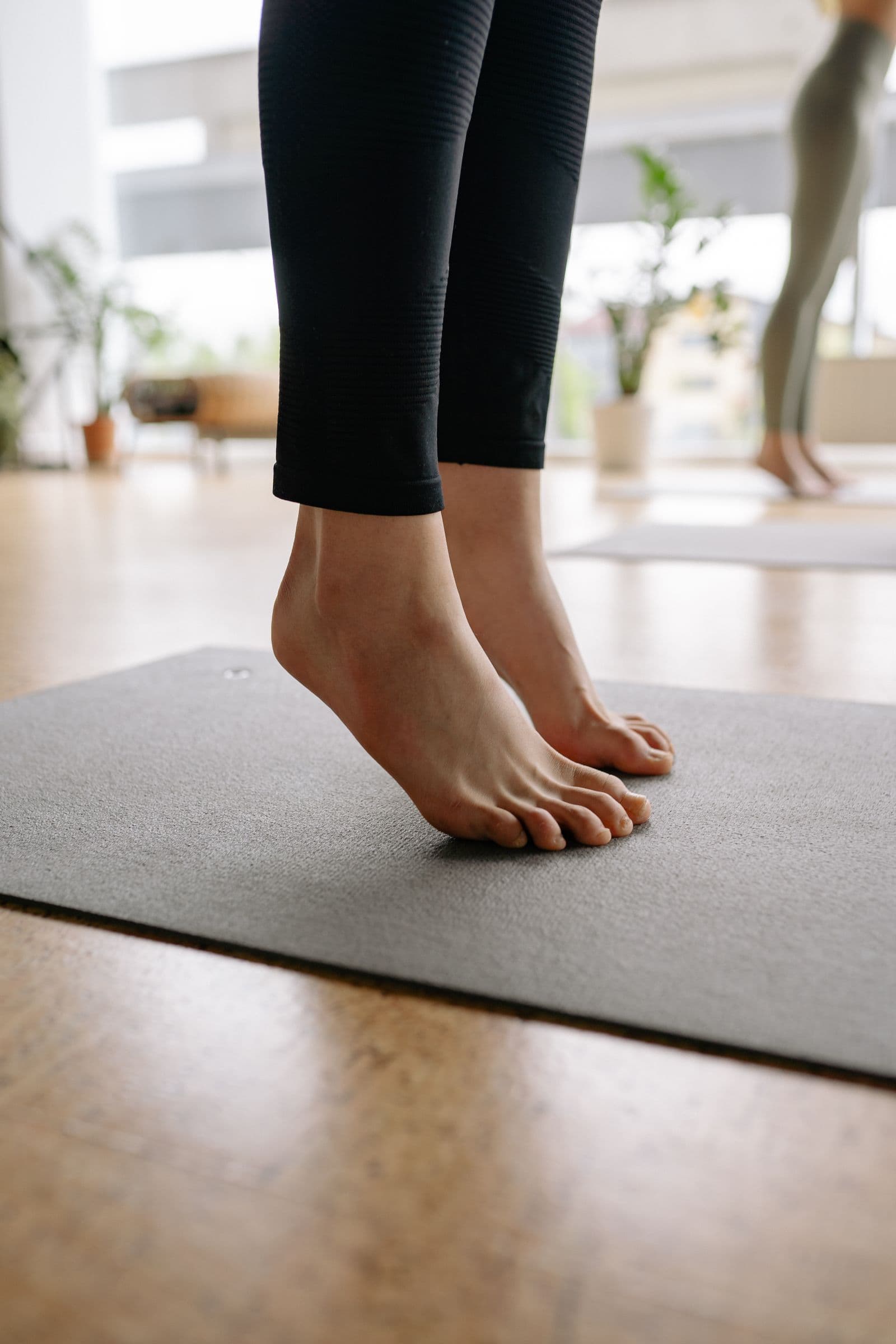 Bare feet rising onto the balls of the feet on a gray yoga mat in a bright room, legs in black leggings
