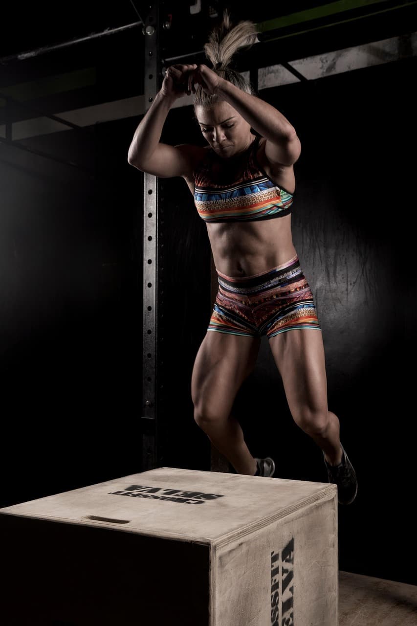 Woman in mid-air above a wooden plyo box during a box jump in a dark gym