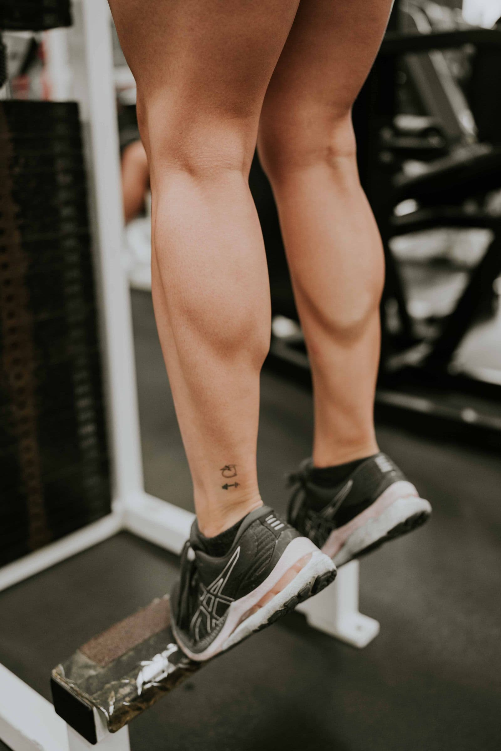 Close-up of calves and running shoes on the edge of a weight bench in a gym, heels raised in a calf raise