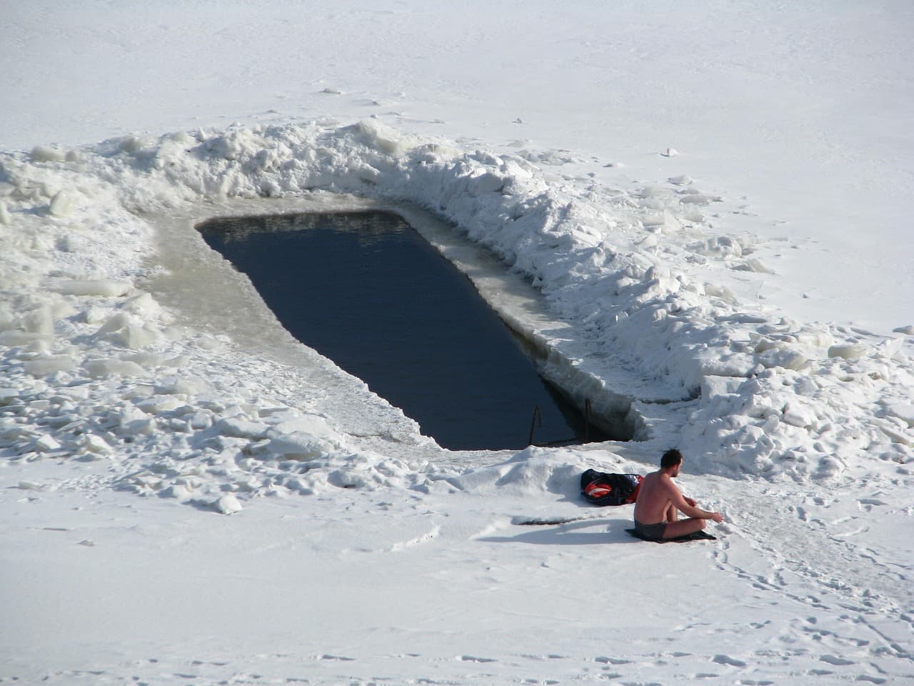 A person in shorts sitting on packed snow beside a rectangular hole cut into a frozen lake for cold water immersion