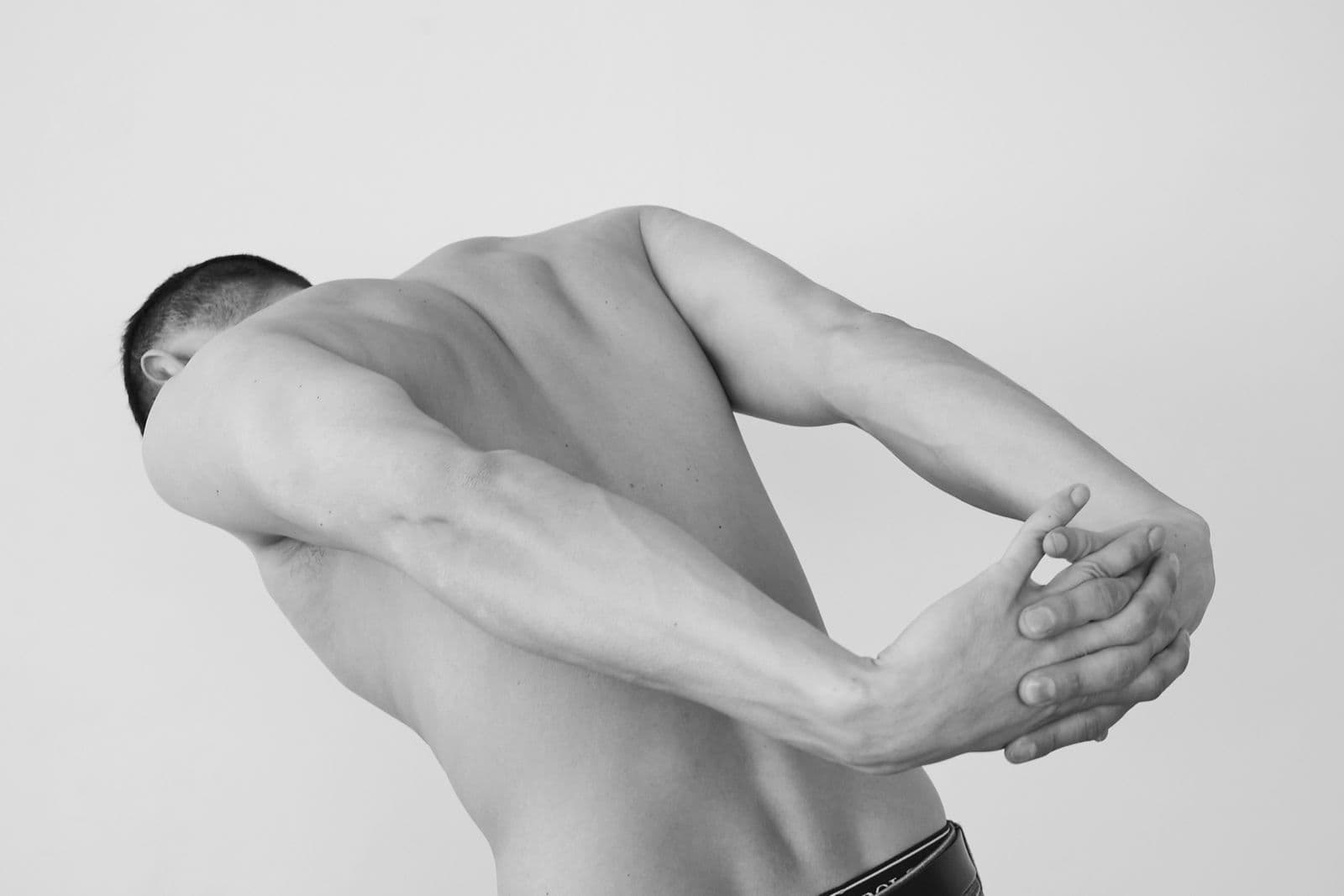 Back view of a person in black and white reaching one arm behind their back to clasp the other at the mid-back, demonstrating shoulder internal rotation reach