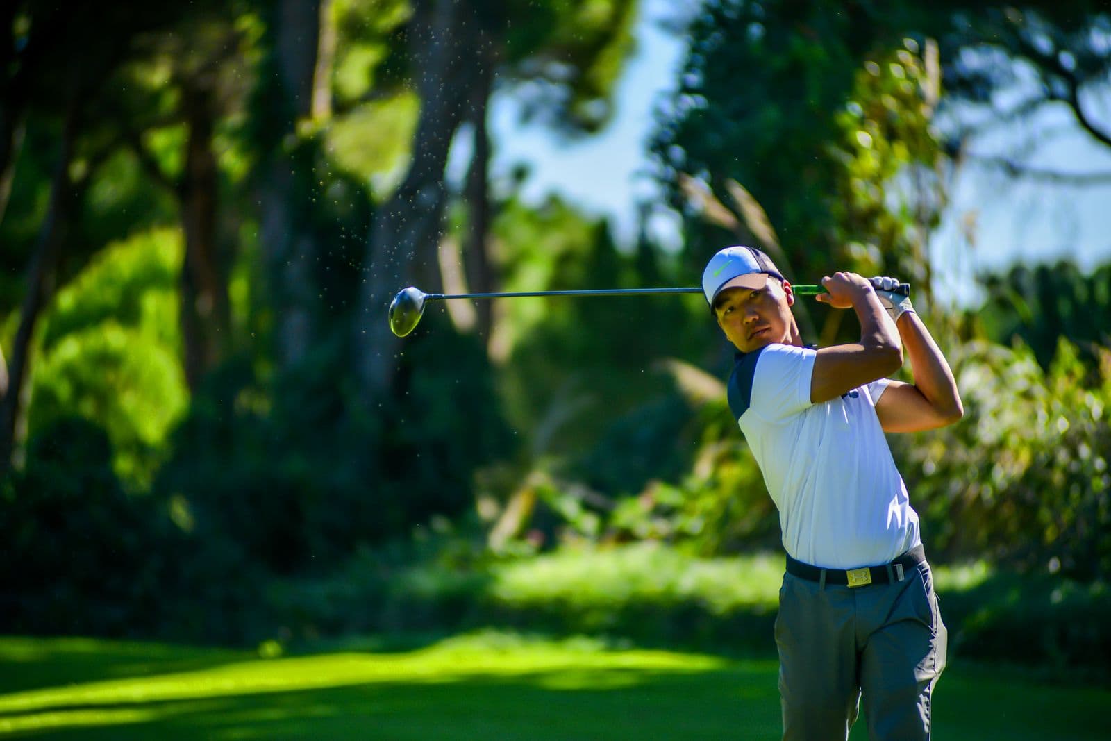 Golfer mid-swing follow-through with club and ball in motion against a green backdrop