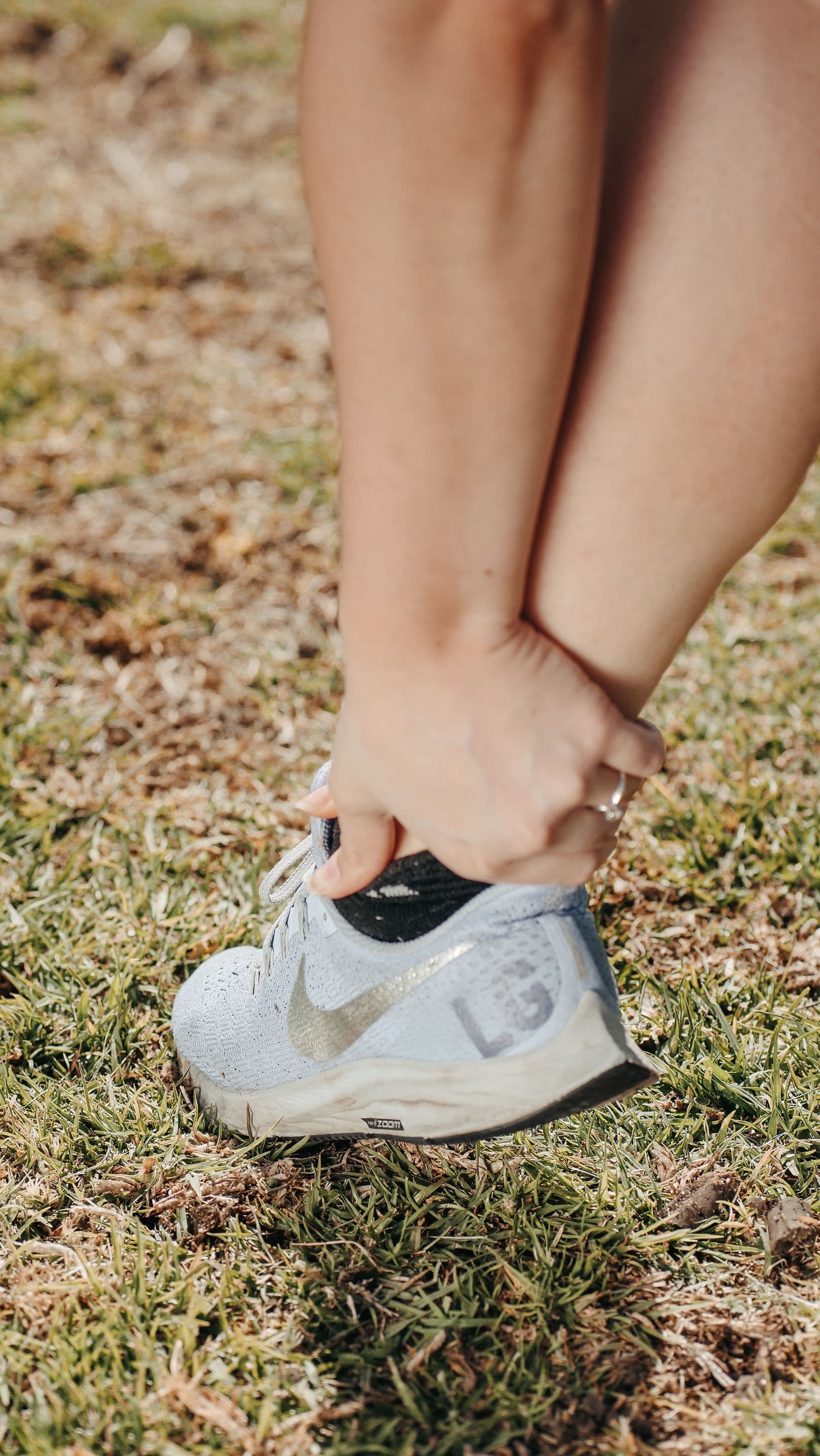 Close-up of a hand gripping the back of a lower leg just above a pale blue running shoe, standing on dry grass