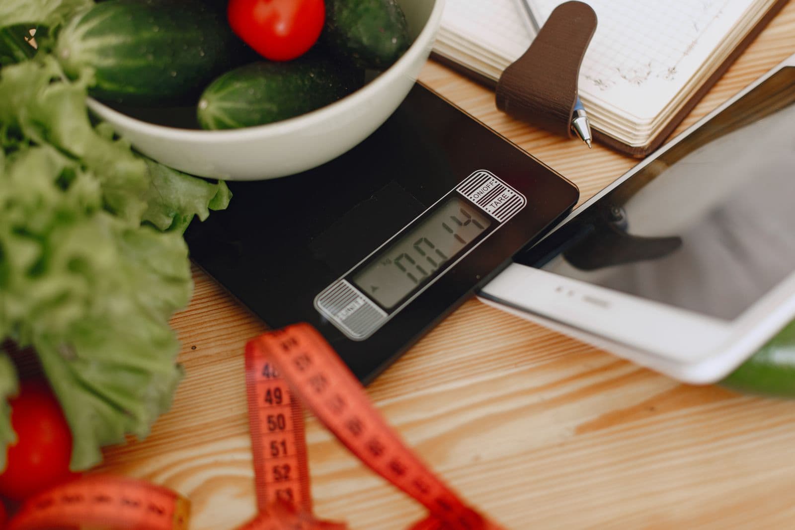 Digital kitchen scale on a wooden counter with a bowl of vegetables, a tape measure, and an open notebook
