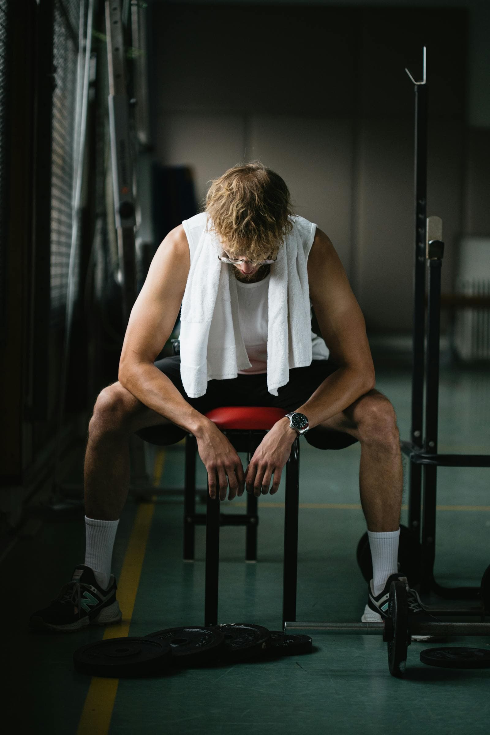 A lifter sitting hunched on a gym bench with a towel around his neck and weight plates on the floor
