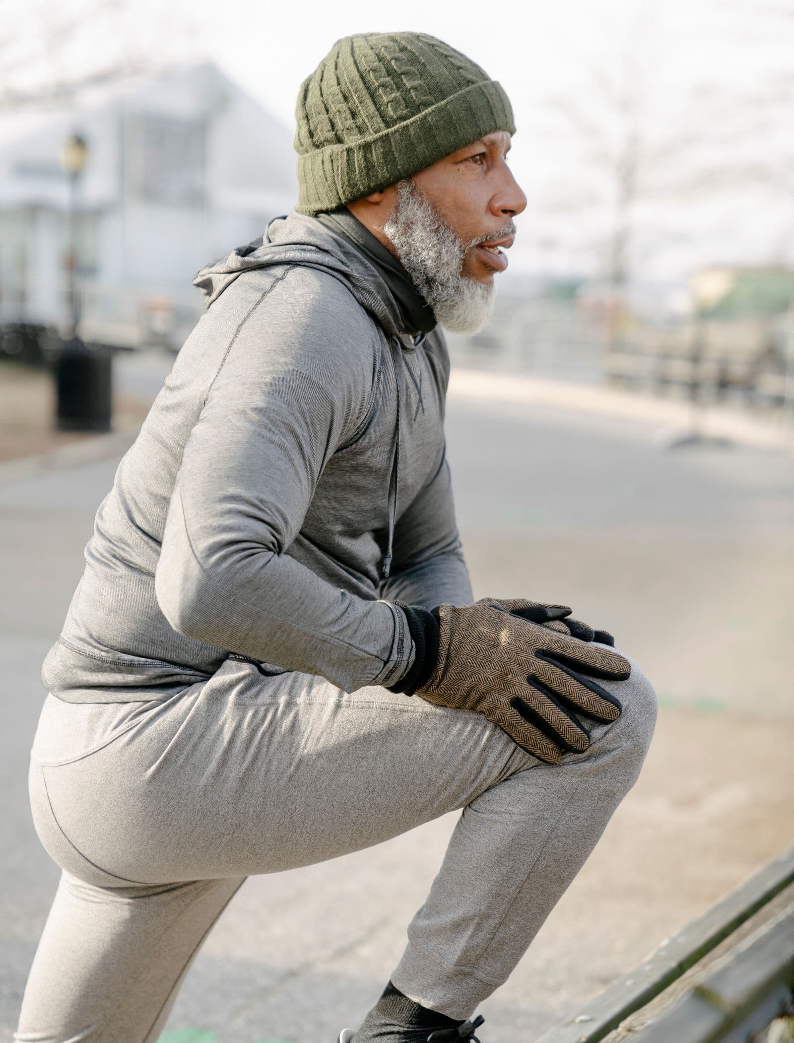 Older man in running clothes and a knit hat holding a lunge stretch outdoors on a cold day