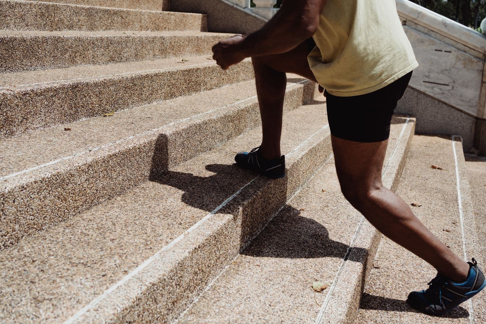 Close view of a person's legs in athletic shoes stepping up a wide outdoor stone staircase in bright sunlight