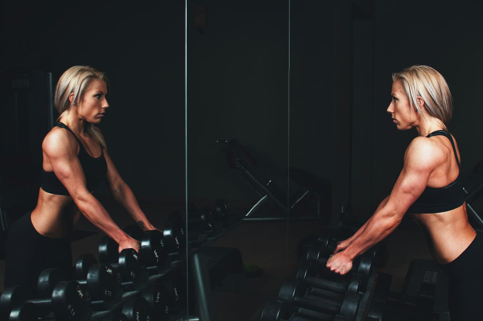 Woman selecting a dumbbell from a rack in a dimly lit gym, mirror reflection visible