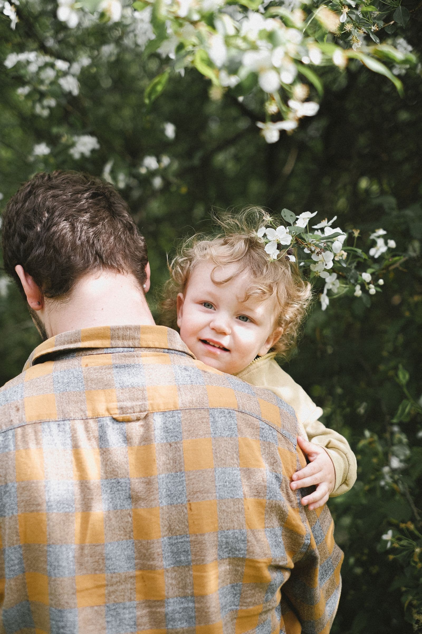A father carrying his toddler over one shoulder in a garden, the child looking back over his back