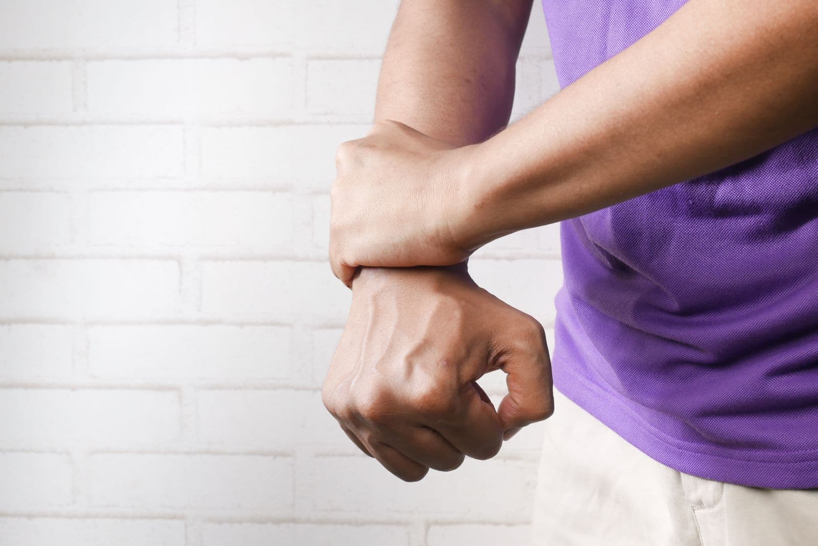 A person in a purple t-shirt gripping one wrist with the other hand in front of a white brick wall