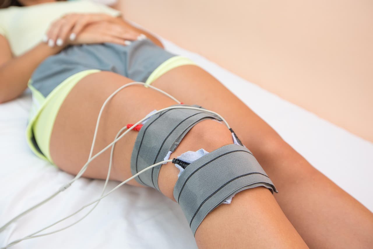Knee wrapped in electrotherapy pads with electrode wires on a clinical table