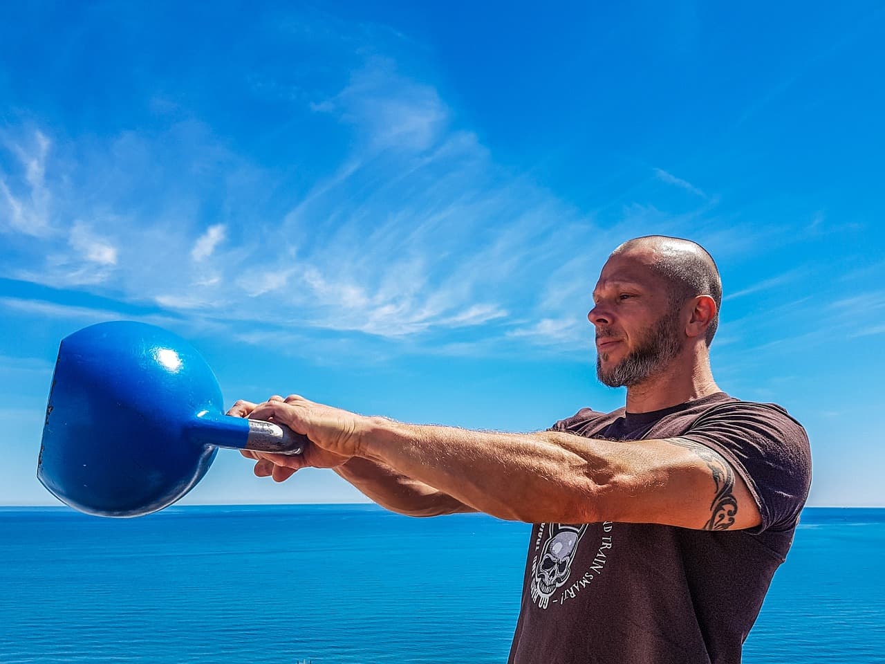A man swinging a blue kettlebell to chest height with both arms extended, ocean and sky behind him