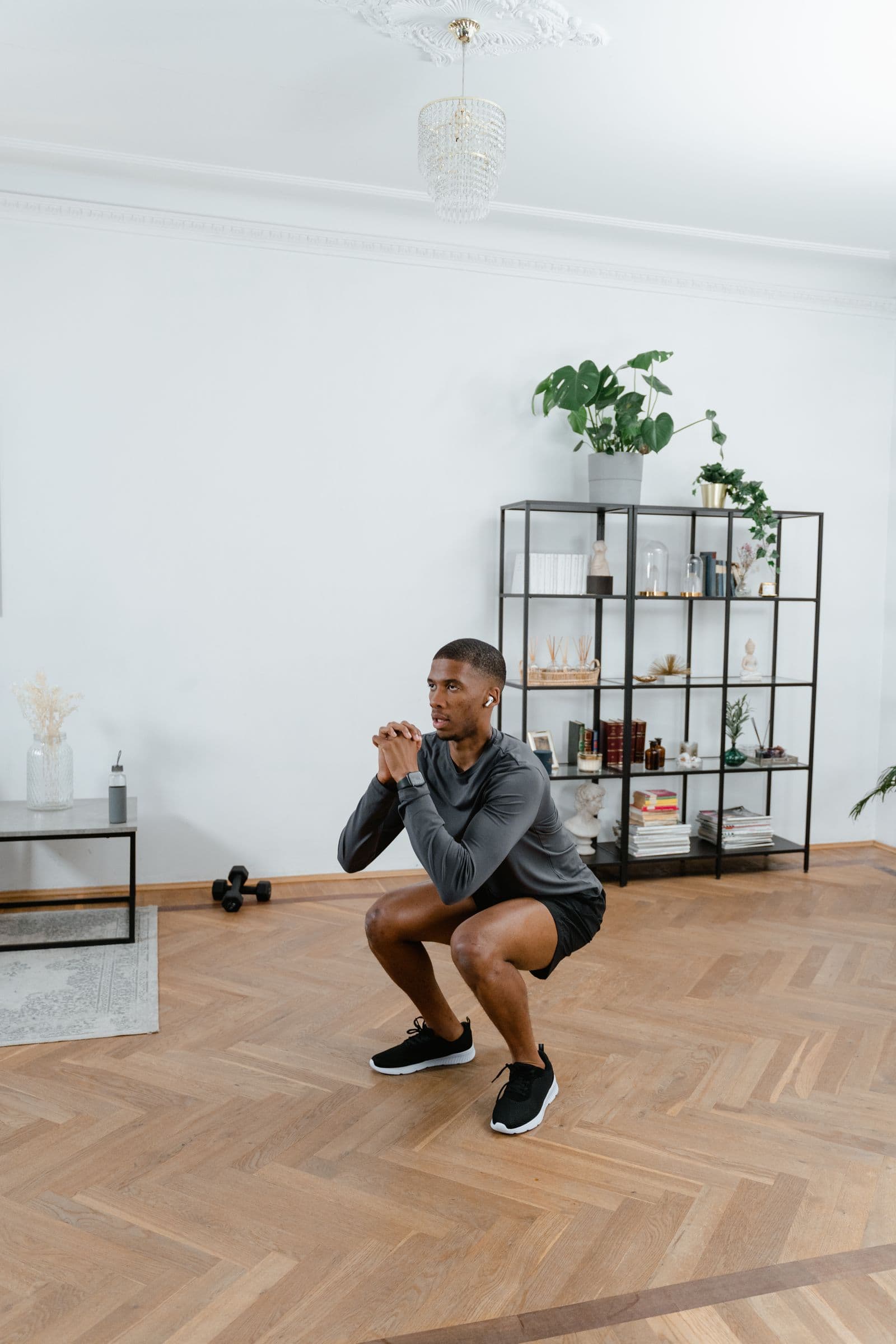 A man in a living room holding the bottom of a controlled bodyweight squat, hands clasped at his chest, dumbbells on the floor behind him