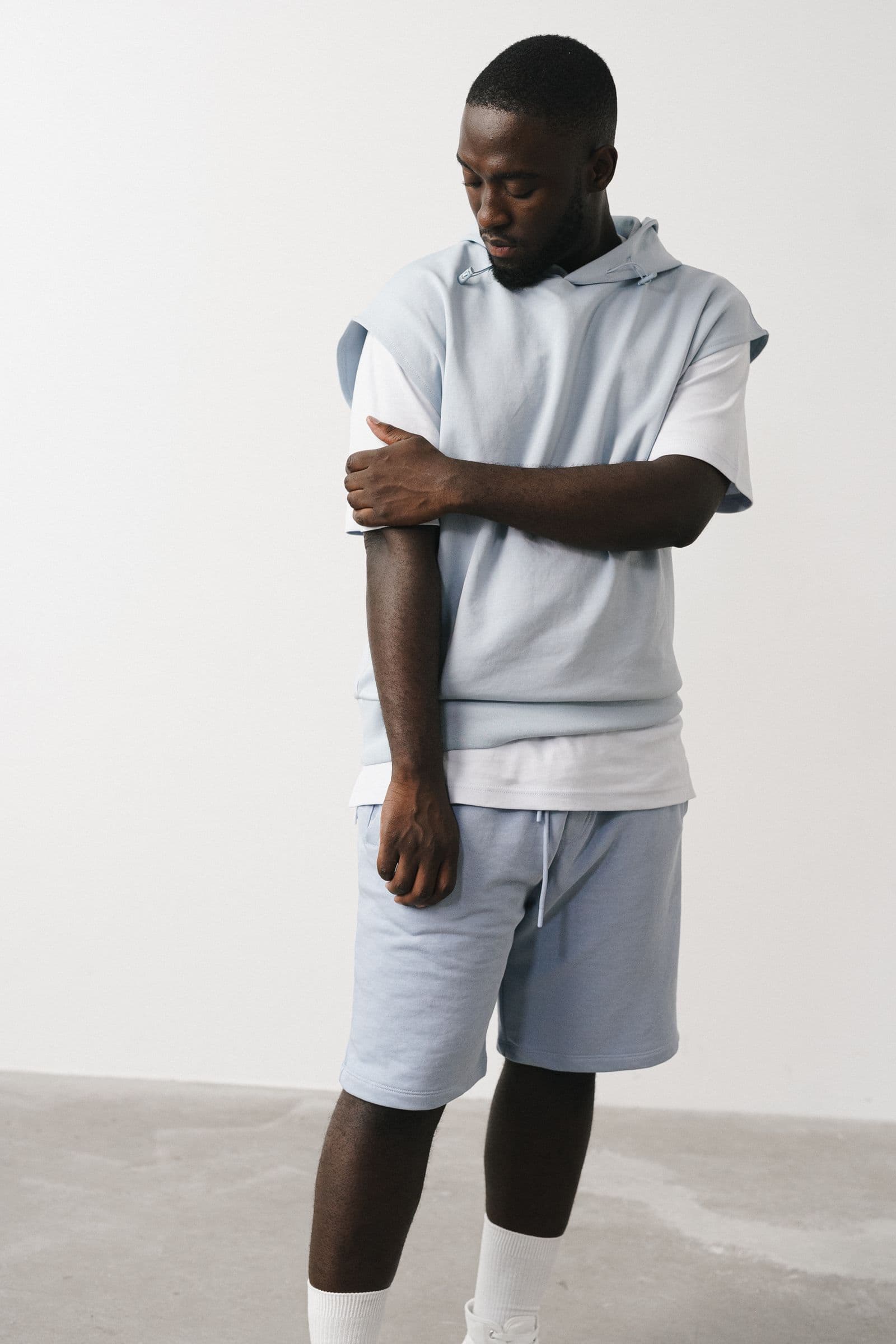 Man in a light blue hoodie and shorts standing against a plain wall, gripping his own forearm just below the elbow and looking down at it