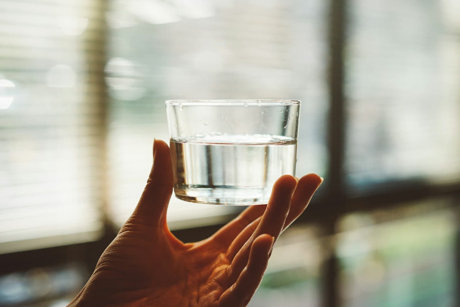 Hand holding a small glass of water by a sunlit window