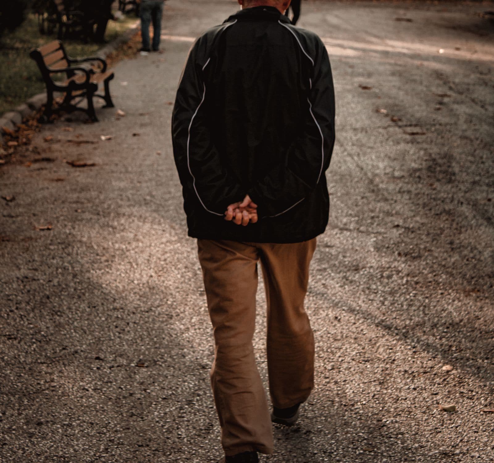 An older man in a dark jacket and khaki trousers walking away down a tree-lined park path with his hands clasped behind his back