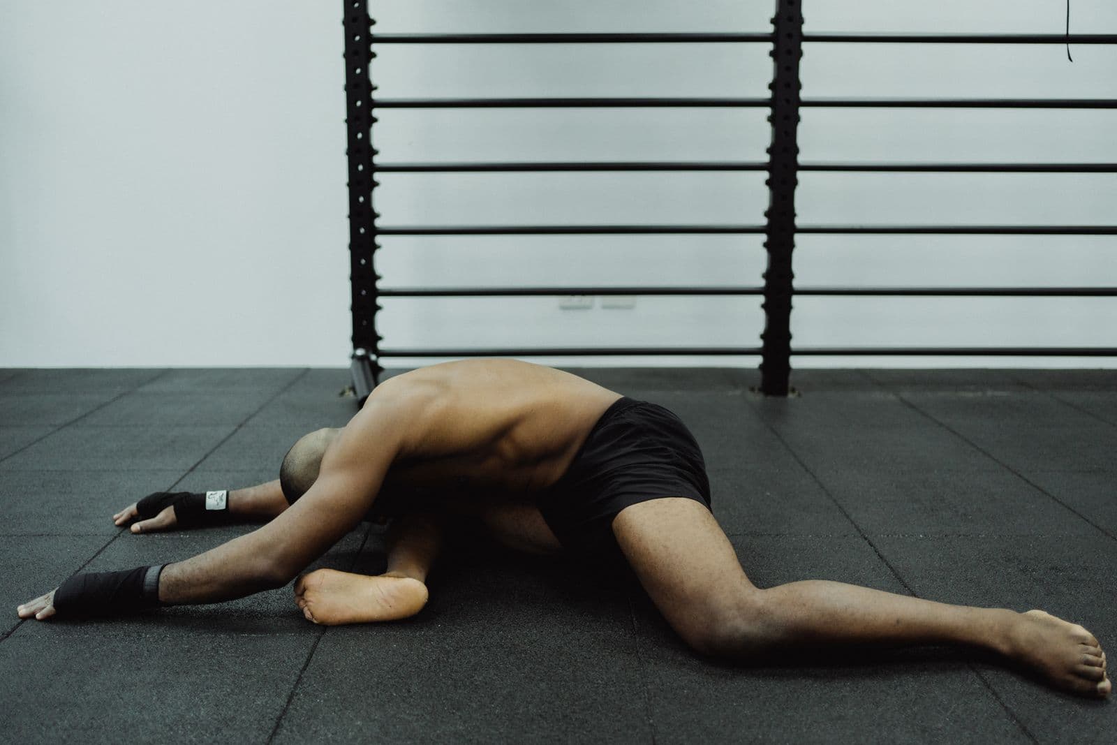 A shirtless man in black shorts folded forward over his front leg in a deep pigeon stretch on a black rubber gym floor, arms reaching ahead