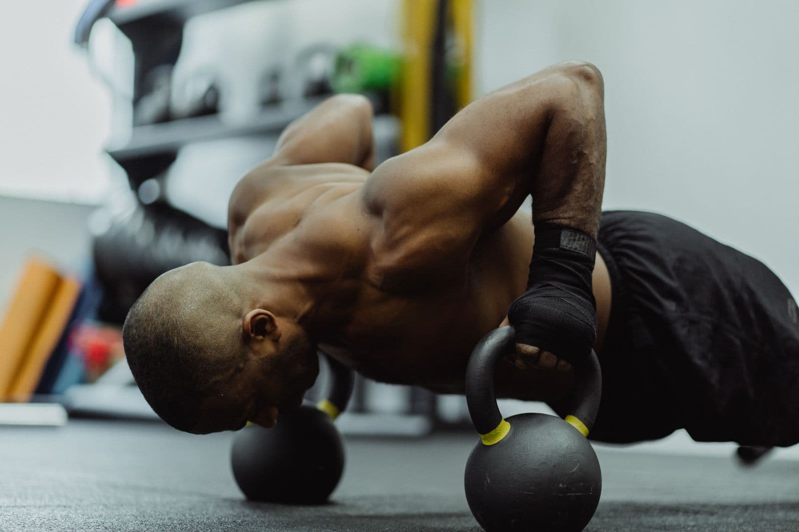 A man in a gym lowering into a push-up with each hand gripping the handle of a kettlebell on the floor, wrists straight