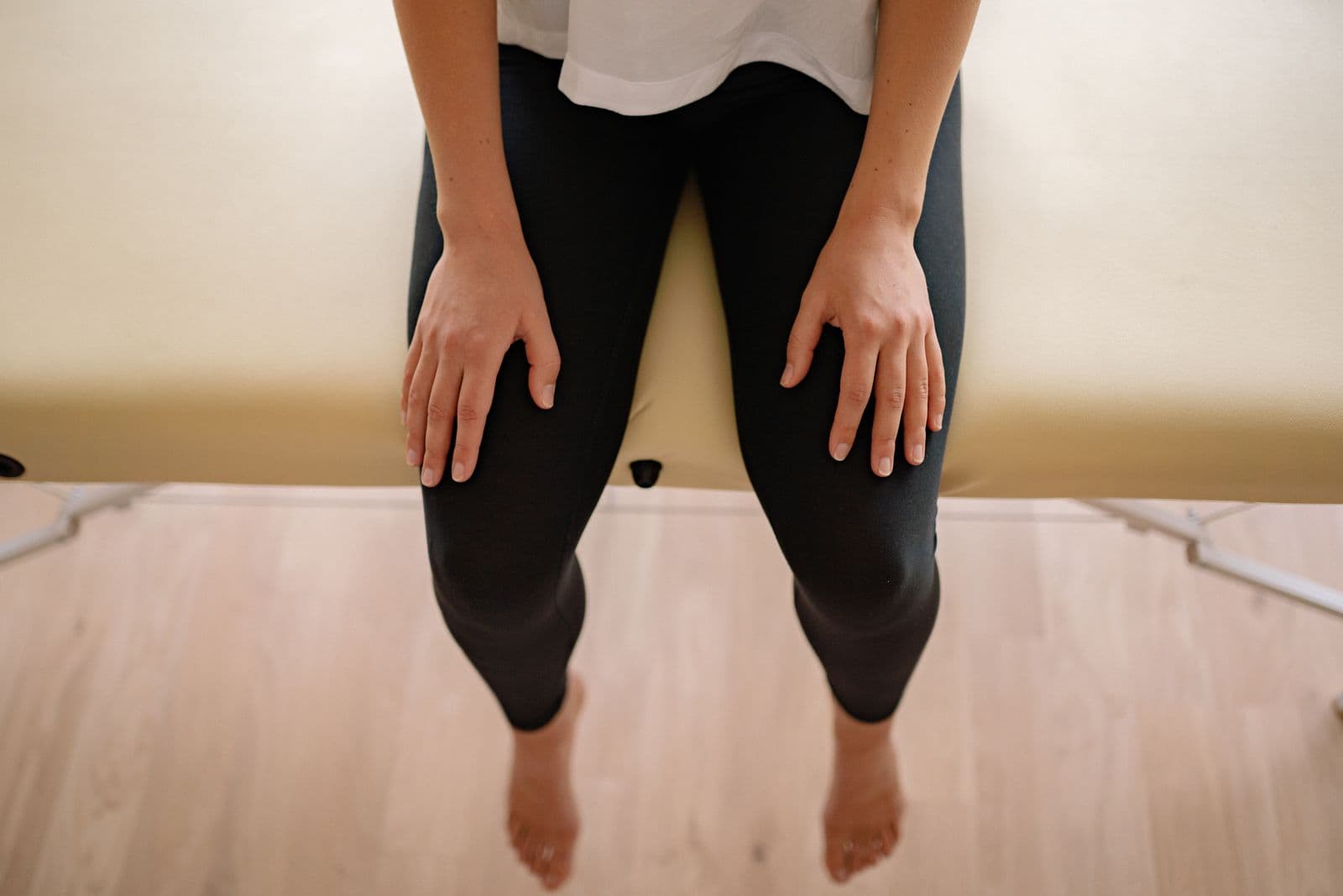 A person in black leggings sitting on the edge of a padded treatment table with bare feet hanging toward a wooden floor, hands resting on their thighs