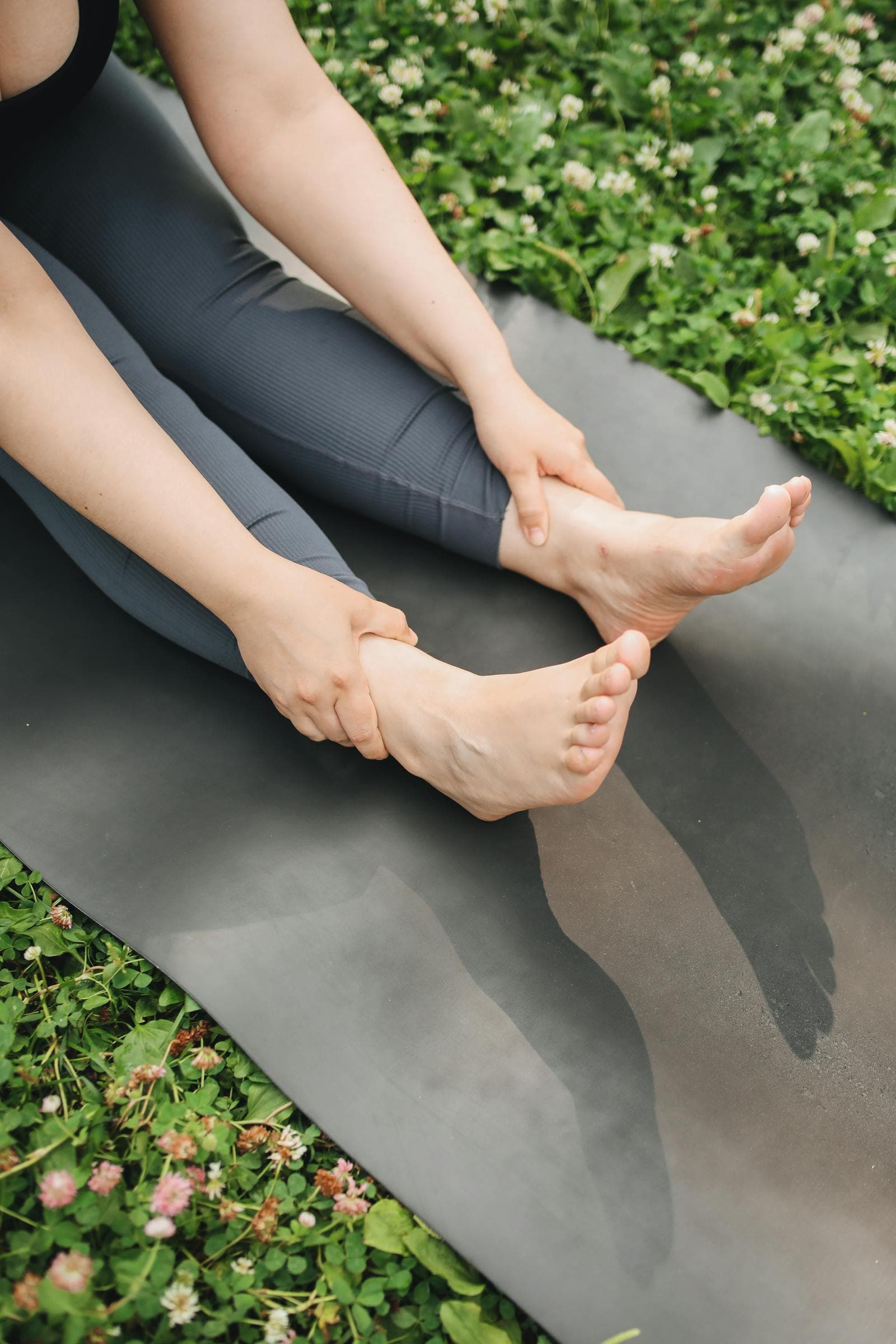 Person sitting on a yoga mat outdoors holding their foot and ankle